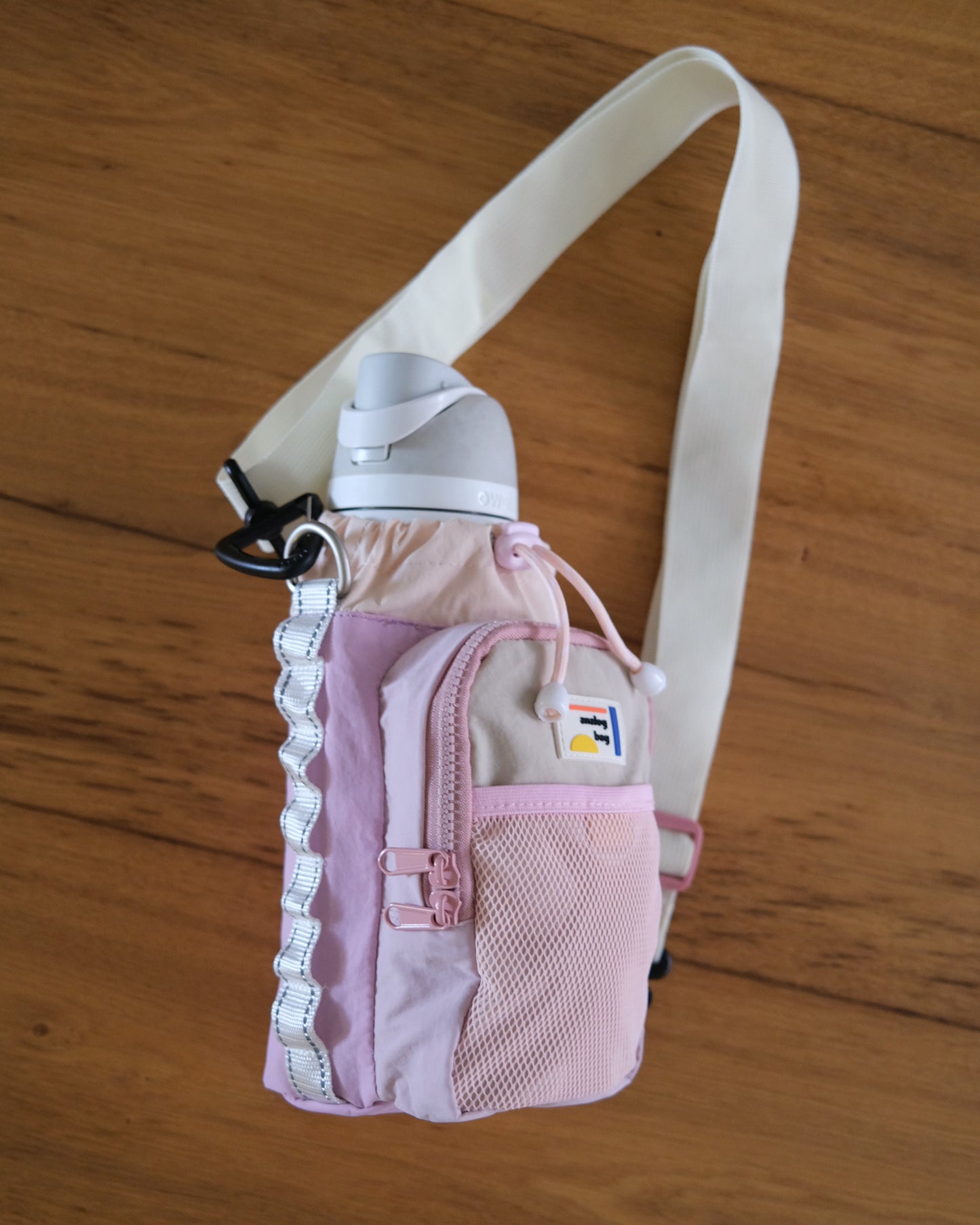 A light pink and lavender water bottle bag with a white shoulder strap, shown on a wooden floor. The bag features a drawstring top, a front zippered pocket, a mesh pocket, and a black carabiner clip.